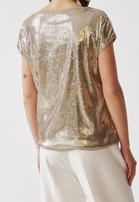 Haut en sequins dorés avec une coupe décontractée et des manches courtes. Présente une surface brillante et texturée ainsi qu'un col rond, associé à un pantalon beige.
