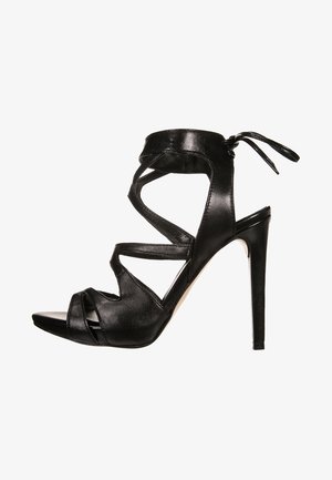 Sandalia negra de tacón alto con tiras de cuero cruzadas, punta abierta y lazo en el tobillo. Presenta un diseño elegante y una superficie texturizada.