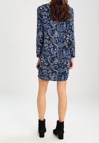 Kleid mit navyblauem Blumenmuster, langen Ärmeln, geradem Schnitt und Rundhalsausschnitt. Trägt dazu schwarze Knöchelstiefel. Weiche Stofftextur.