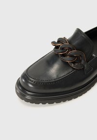 Mocassin en cuir noir avec un accent de chaîne épais en noir et bronze. Dispose d'un bout rond et d'une semelle en caoutchouc texturée pour une meilleure adhérence.