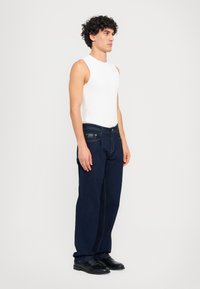 Temno modri jeans s ravnimi nogavicami, ki imajo pet žepov in kontrastno rumeno šivanje, v kombinaciji z belo brezrokavno majico.