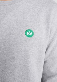 Felpa grigia in misto cotone, con un logo ricamato verde circolare e un design bianco sul lato sinistro del petto. Texture liscia.