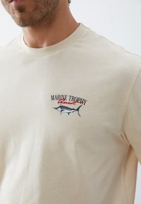 Beige cotton t-shirt with a round neck, featuring a colorful embroidered logo of a fish and text “MARINE TROPHY.” Smooth texture.