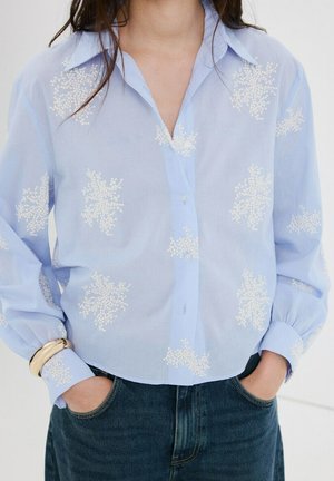 Mujer con blusa de botones azul claro con bordado floral blanco, manos en los bolsillos de pantalones vaqueros azul oscuro y pulsera dorada en la muñeca izquierda.