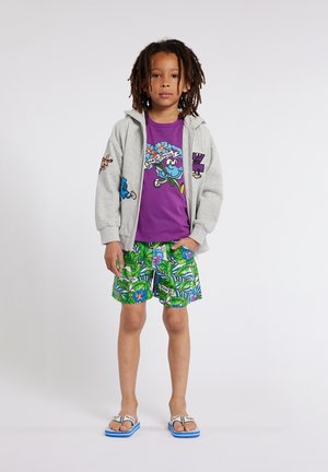 Felpa grigia con zip e grafiche colorate, t-shirt viola con design floreale, pantaloni corti verdi con motivo tropicale e infradito con suola blu.