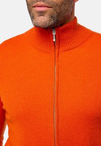 Pull de couleur orange zippé avec un col montant, présentant une texture côtelée et une fermeture éclair argentée. La vue rapprochée met en avant la douceur du tissu.