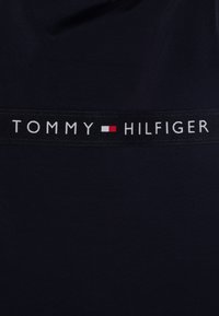 Marine tekstil med et svart bånd som har "TOMMY HILFIGER" i hvit tekst, akkompagnert av et rødt og hvitt rektangel. Glatt tekstur.