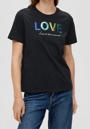 Kobieta ubrana w czarną koszulkę z cekinowym, tęczowym napisem "LOVE" oraz białym, kursywnym sloganem "Love is the answer", w połączeniu z niebieskimi dżinsami.