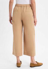 Helle beige weit geschnittene Hose mit elastischem Bund, zwei Gesäßtaschen und glatter Textur. Kombiniert mit braunen Slippern.