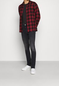 Camisa de franela a cuadros rojos y negros, de manga larga, camiseta negra debajo, jeans oscuros, zapatillas blancas, pose de pie, textura suave.
