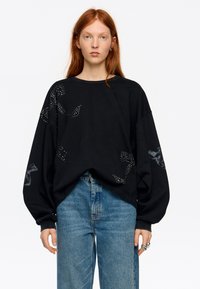 Sudadera negra de gran tamaño con mangas abullonadas, con adornos que parecen flores en tachuelas plateadas. Llevada sobre vaqueros de mezclilla azul claro.