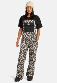 Kobieta w beżowej, dzianinowej czapce, czarnej koszulce, ogrodniczkach w czarno-białe wzory w panterkę z przednią kieszenią oraz czarnych trampkach stoi na tle białym.