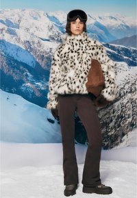 Faux fur jacket with black spots over white, paired with brown pants and gloves. Model wears ski goggles against a snowy mountain backdrop.