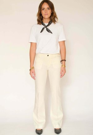 T-shirt blanc, pantalon évasé crème et chaussures noires à motifs. La tenue est complétée par un foulard noir noué autour du cou et des détails en tissu texturé.