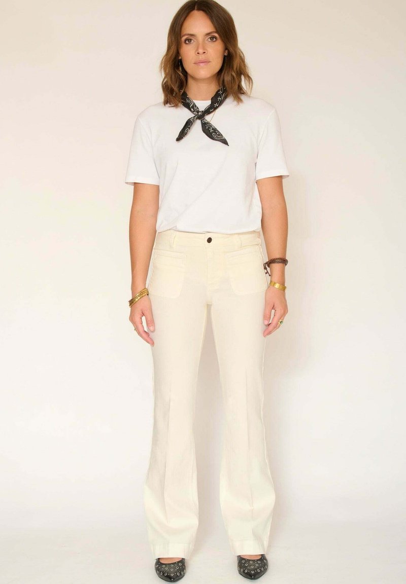 T-shirt blanc, pantalon évasé crème et chaussures noires à motifs. La tenue est complétée par un foulard noir noué autour du cou et des détails en tissu texturé.