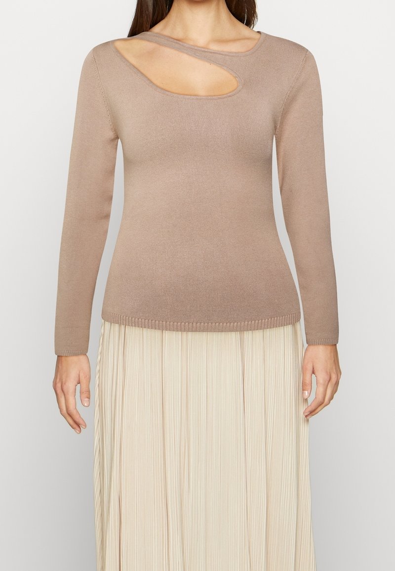 Femme portant un pull beige à manches longues avec une découpe asymétrique au niveau du col et une jupe plissée crème.