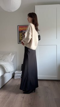 Υπερμεγέθη ζακέτα από κρεμ πλεκτό, μαύρη φούστα μήκους έως το πάτωμα και μαύρες μπότες με τακούνι. Δωμάτιο με ξύλινο πάτωμα και ορατό λευκό κρεβάτι στο βάθος.