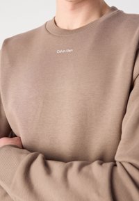 Personne portant un sweat-shirt beige Calvin Klein avec poignets côtelés et col rond, bras croisés sur la poitrine.