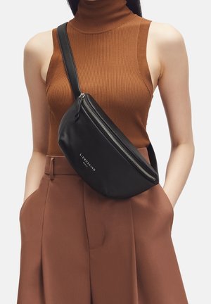 Černá kožená crossbody taška s zakřiveným tvarem, stříbrným zipem a embosovaným logem. Nositelná přes hnědý žebrovaný rolák.
