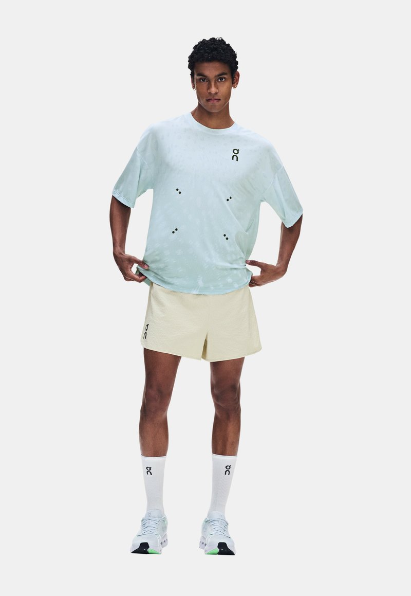 Homme en t-shirt bleu clair et short beige, portant des chaussettes blanches et des baskets, debout les mains sur les hanches sur un fond uni.