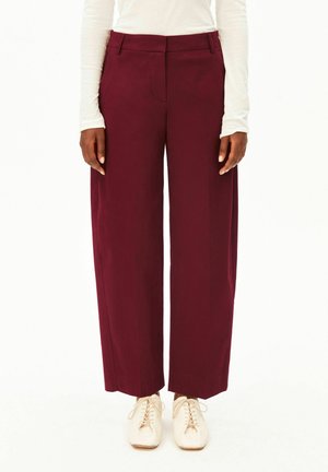 Femme portant un pantalon bordeaux taille haute et jambes larges, associé à des chaussures blanches à lacets et un haut blanc à manches longues, debout, les bras le long du corps.
