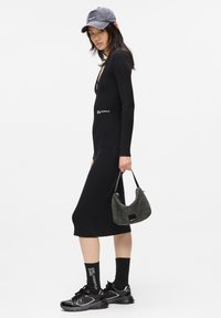 Kobieta w czarnej, prążkowanej sukience midi, czarnej czapce, skarpetkach z białym napisem, czarnych sneakersach, trzymająca szary, puszysty torebkę na tle białym.