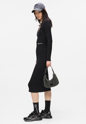 Mujer con vestido midi negro de canalé, gorra negra, calcetines con texto blanco, zapatillas negras, sosteniendo un bolso de felpa gris contra un fondo blanco.