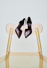 Escarpins slingback à bout pointu marron foncé reposant sur le dossier d'une chaise en plastique beige translucide, devant un mur clair.