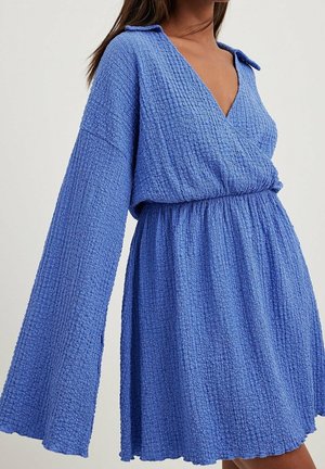 Kvinde iført en blå tekstureret langærmet kjole med dyb V-hals og indsnævret talje, vist fra skuldrene til midt på låret.