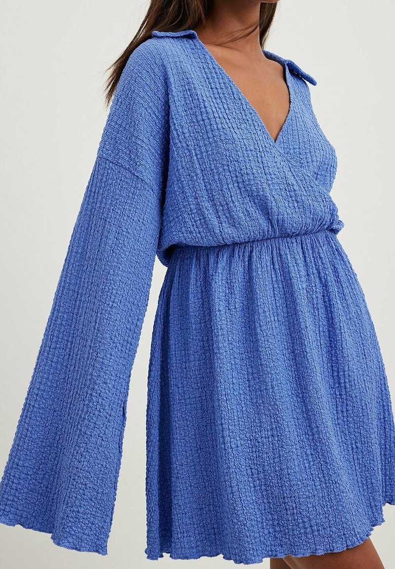 Femme portant une robe bleue texturée à manches longues avec un col en V profond et une taille froncée, montrée des épaules jusqu'à mi-cuisse.