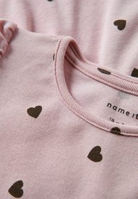 Haut en coton rose avec un col rond à côtes orné de petits motifs en forme de cœur marron. Comprend une étiquette de soin près du col.