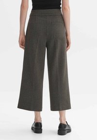 Hnědé kalhoty s kostkovaným vzorem ve stylu houndstooth, široké nohavice s vysokým pasem a zadní kapsou, vyrobené z texturované látky, doplněné černými botami.