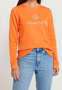 Felpa arancione in cotone, con scollatura rotonda e polsini a coste. Logo "GANT" bianco ben visibile sulla parte frontale.