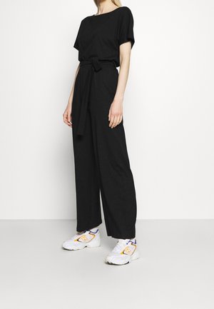 Sort jumpsuit med afslappet pasform, korte ærmer og knyttet taljebælte. Kombineret med hvide sneakers med orange og lilla detaljer.
