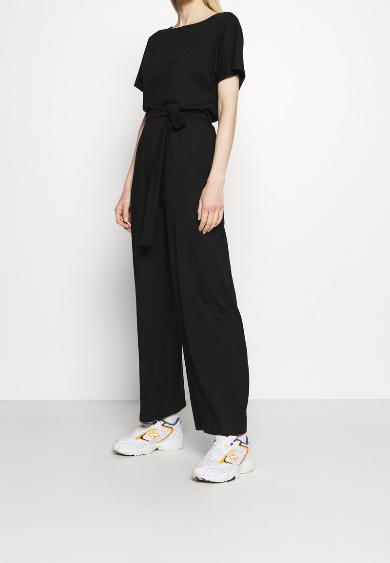 Combinaison noire à coupe décontractée, manches courtes et ceinture nouée à la taille. Associée à des baskets blanches avec des touches orange et violettes.