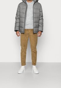 Uomo che indossa un giubbotto imbottito grigio, un maglione grigio chiaro, pantaloni in velluto a coste marroni e sneakers bianche, in piedi contro un muro bianco uniforme.
