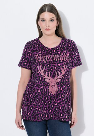 Frau mit glattem braunem Haar trägt ein lila-schwarzes Leopardenmuster-T-Shirt mit einem rosa glitzernden Hirschkopf und dem Wort „Herzmadi“ in Glitzerschrift.