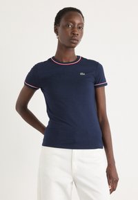 T-shirt bleu marine en coton doux, avec des accents de col et de manches en ribbing rouge et blanc. Petit logo vert sur la poitrine.