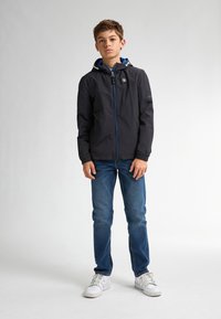 Jachetă ușoară neagră cu glugă albastră, fermoar frontal și detalii cu logo. Purtați cu blugi albaștri și adidași albi, stând în fața unui fundal neutru.