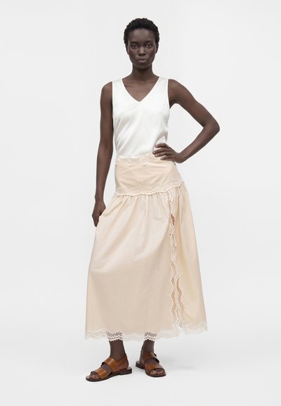 Femme portant un haut en satin blanc sans manches, une longue jupe bordée de dentelle crème et des sandales en cuir marron, debout, une main sur la hanche.