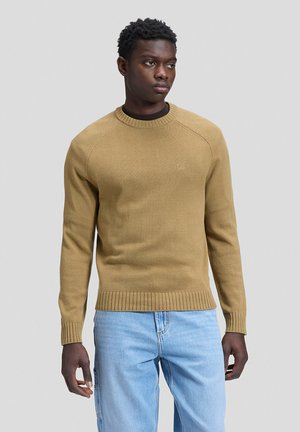Ein Mann steht nach vorne gerichtet und trägt einen beigefarbenen Strickpullover und hellblaue Jeans vor einem einfarbigen, hellgrauen Hintergrund. Der Gesichtsausdruck ist neutral.