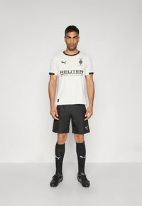 Fehér futballmez fekete díszítésekkel és REUTER logóval, fekete rövidnadrággal és zoknival párosítva. A fekete stoplis cipő egészíti ki a sportos megjelenést.