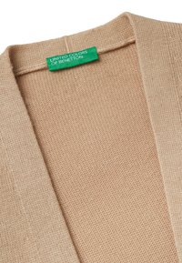 Beige pletený cardigan s texturovaným povrchem, volným předním designem a zelenou značkovou nálepkou s nápisem "United Colors of Benetton."