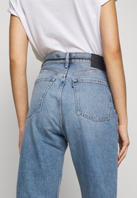 Světle modré džíny s vysokým pasem, které mají jednobarevný zadní díl, dvě zadní kapsy a koženou etiketu na pasu. Vyrobeno z denimového materiálu.