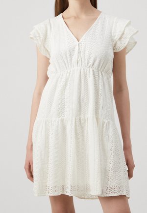 Mujer con un vestido blanco de encaje con escote en V, mangas amplias y cintura fruncida, posando frente a un fondo liso y claro.
