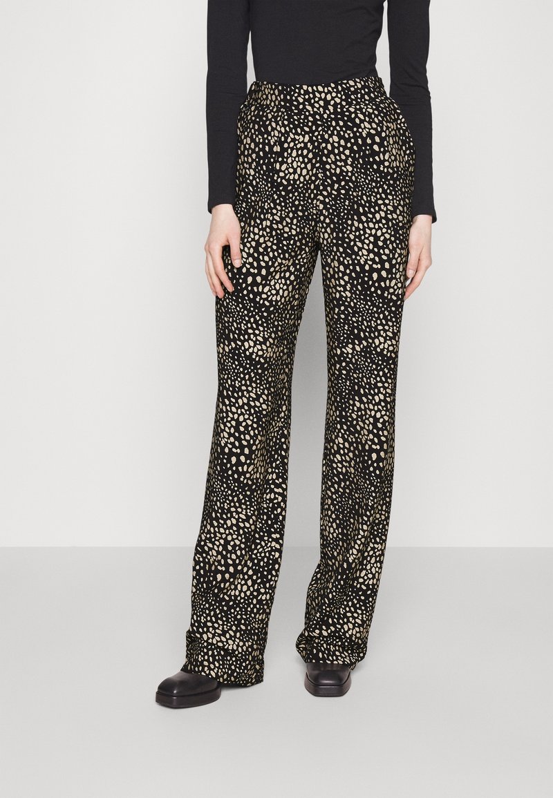 Femme portant un pantalon large noir à motif beige tacheté, associé à un haut noir et des chaussures noires, se tenant devant un fond uni.