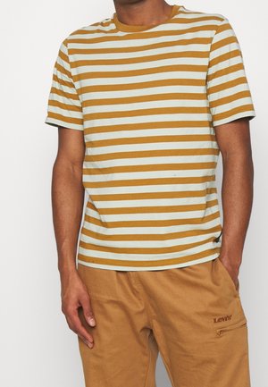 Homme portant une chemise à manches courtes à rayures horizontales moutarde et crème, associée à un pantalon Levi's beige avec poche zippée.