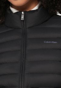 Κοντινό πλάνο ενός ατόμου που φοράει μια μαύρη επενδυμένη ζακέτα με φερμουάρ και ψηλό γιακά, με το λογότυπο της Calvin Klein στο στήθος.