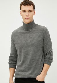 Suéter de cuello alto gris en punto texturizado, con mangas largas y un ajuste ceñido, llevado con una postura relajada y las manos en los bolsillos.