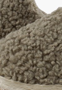 Le pantofole in shearling beige presentano una superficie testurizzata con un aspetto morbido e riccio. La suola è rivestita con un materiale chiaro in contrasto.